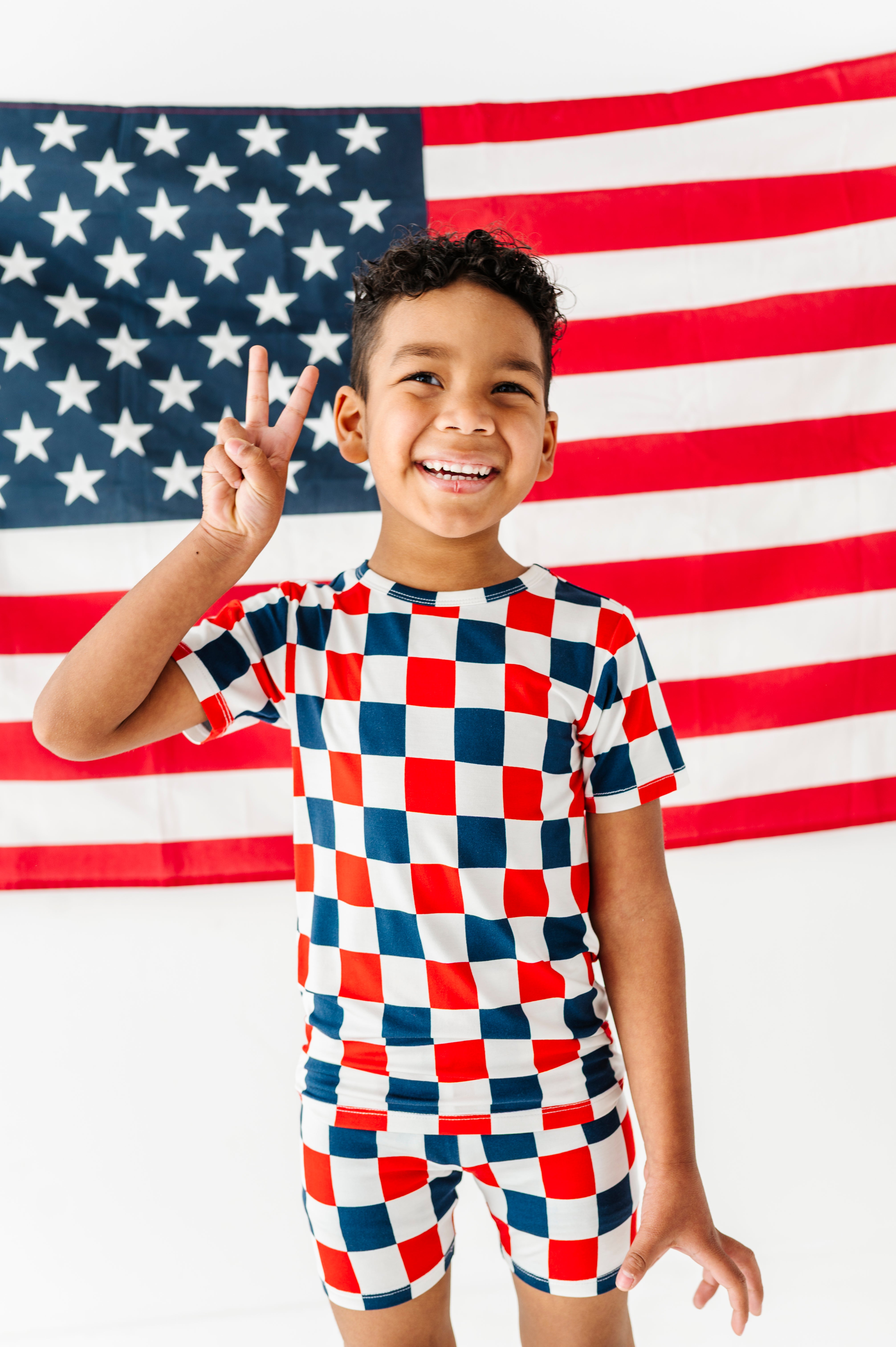 Child wearing a red, white, and blue checkered bamboo two-piece pajama outfit in front of an American flag